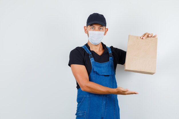 Jeune chargeur montrant un sac en papier brun en uniforme, masque, vue de face.