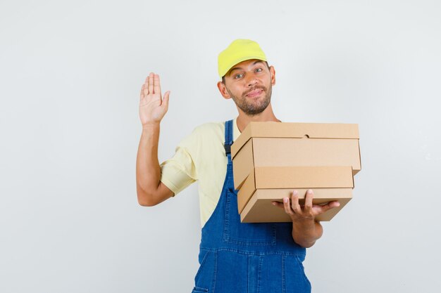 Jeune chargeur en uniforme tenant des boîtes en carton et disant bonjour, vue de face.