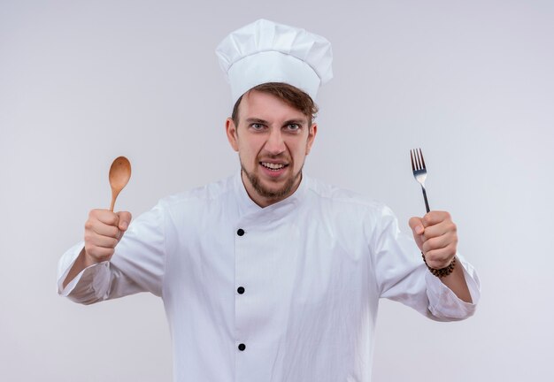 Un jeune chef barbu affamé homme vêtu d'un uniforme de cuisinière blanc et chapeau tenant une cuillère en bois et une fourchette tout en regardant sur un mur blanc