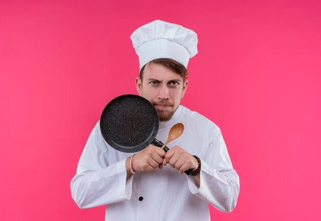 Un jeune chef barbu agressif homme en uniforme blanc montrant x signe avec poêle et cuillère en bois tout en regardant sur un mur rose