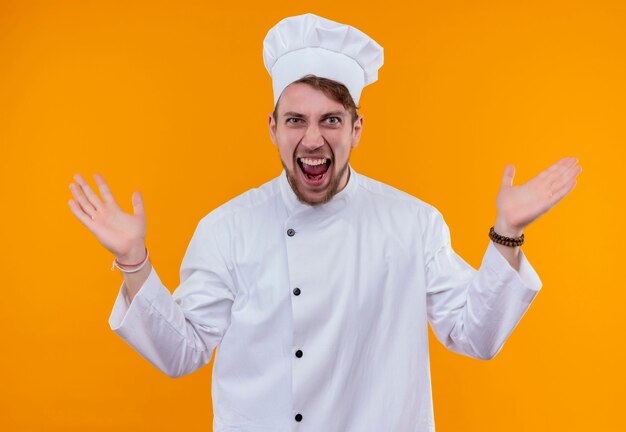 Un jeune chef barbu agressif homme en uniforme blanc ouvrant ses mains tout en regardant sur un mur orange