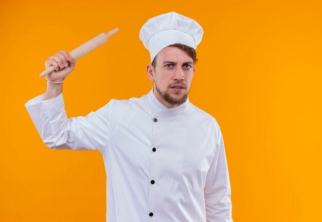 Un jeune chef barbu agressif homme en uniforme blanc soulevant le rouleau à pâtisserie tout en regardant sur un mur orange