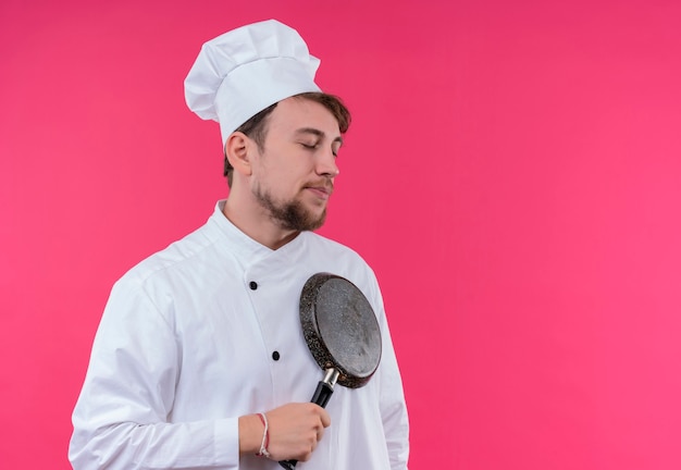 Un jeune chef barbu calme homme en uniforme blanc tenant une poêle à frire avec les yeux fermés sur un mur rose
