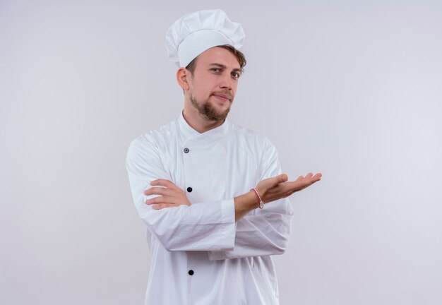 Un jeune chef barbu confiant homme vêtu d'un uniforme de cuisinière blanche et chapeau levant la main et tenant les mains jointes en regardant sur un mur blanc