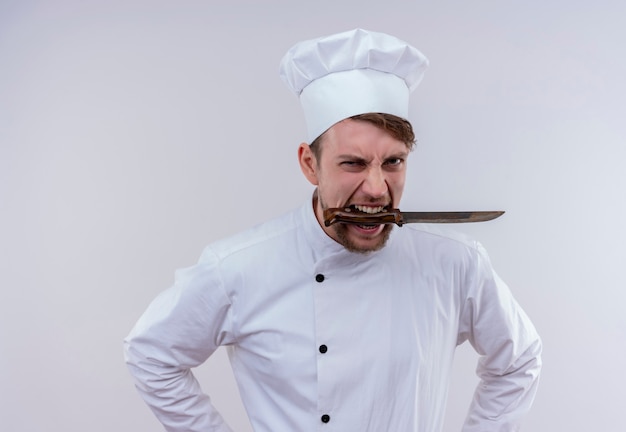 Photo gratuite un jeune chef barbu furieux homme vêtu d'un uniforme de cuisinière blanc et chapeau tenant un couteau sur sa bouche tout en regardant sur un mur blanc