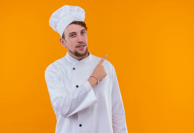 Un jeune chef barbu heureux homme en uniforme blanc pointant vers le haut avec l'index sur un mur orange