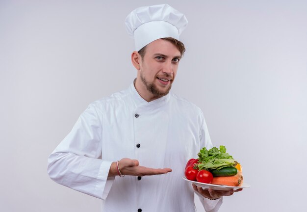 Un jeune chef barbu heureux homme vêtu d'un uniforme de cuisinière blanc et chapeau montrant une assiette blanche avec des légumes frais tels que tomates, concombres, laitue sur un mur blanc