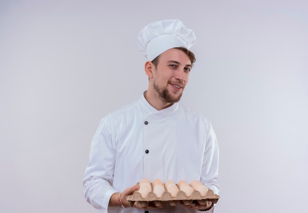 Un jeune chef barbu heureux homme vêtu d'un uniforme de cuisinière blanc et chapeau tenant un carton d'oeufs tout en regardant sur un mur blanc