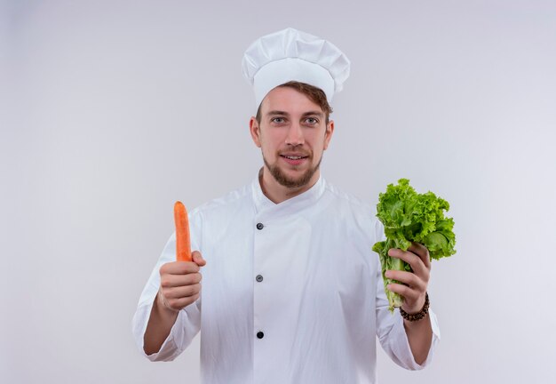 Un jeune chef barbu heureux homme vêtu d'un uniforme de cuisinière blanche et hat holding green leaf lettuce avec carotte tout en regardant sur un mur blanc