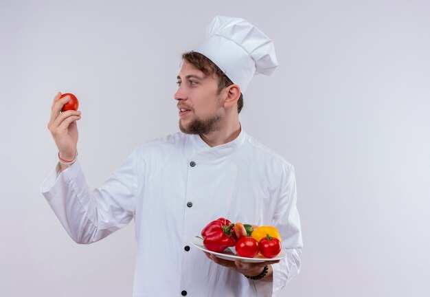 Un jeune chef barbu homme portant l'uniforme de cuisinière blanche et un chapeau à la tomate tout en tenant une assiette blanche avec des légumes frais tels que les tomates, les concombres, la laitue sur un fond blanc