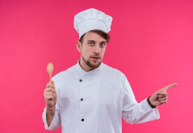 Un jeune chef barbu homme en uniforme blanc côté pointant avec l'index, tenant une cuillère en bois tout en regardant sur un mur rose