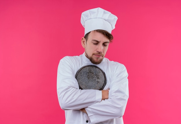 Un jeune chef barbu homme en uniforme blanc hugging poêle à frire avec les yeux fermés sur un mur rose