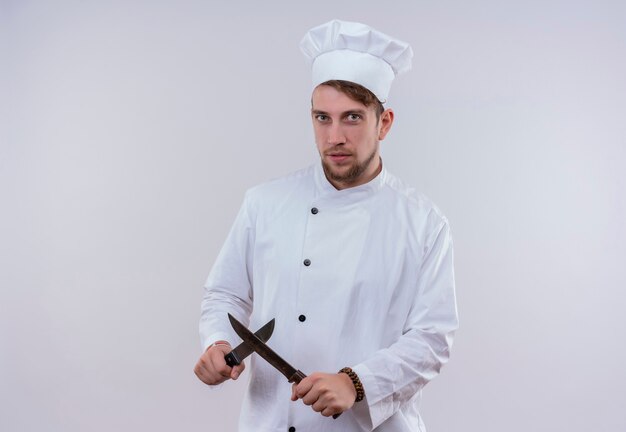 Un jeune chef barbu homme vêtu d'un uniforme de cuisinière blanc et chapeau montrant x signe avec couteau tout en regardant sur un mur blanc