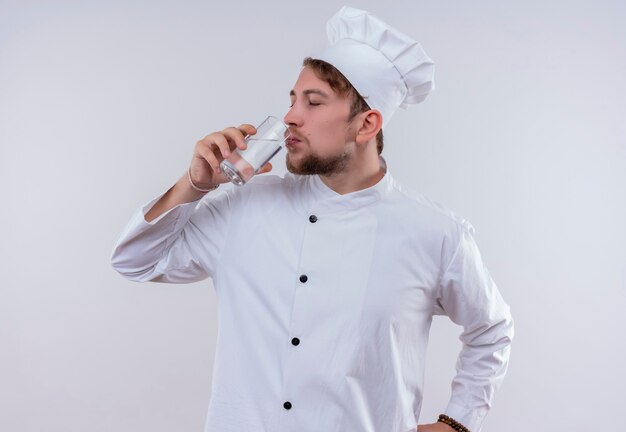 Un jeune chef barbu homme vêtu d'un uniforme de cuisinière blanche et chapeau de boire un verre d'eau tout en regardant sur un mur blanc