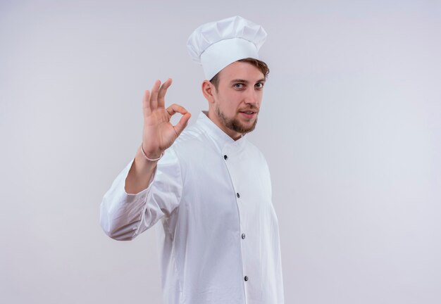 Un jeune chef barbu joyeux homme en uniforme blanc montrant le geste ok sur un mur blanc