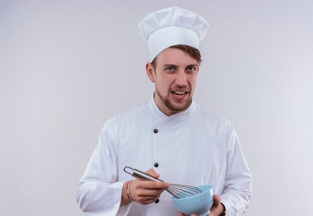 Un jeune chef barbu mécontent homme vêtu d'un uniforme de cuisinière blanc et chapeau tenant un bol bleu avec une cuillère mélangeuse tout en regardant sur un mur blanc