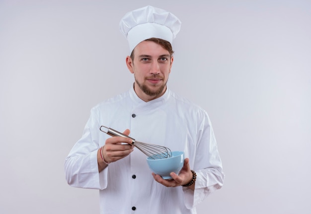 Un jeune chef barbu souriant homme vêtu d'un uniforme de cuisinière blanc et chapeau tenant la cuillère de mixage sur un bol bleu tout en regardant sur un mur blanc