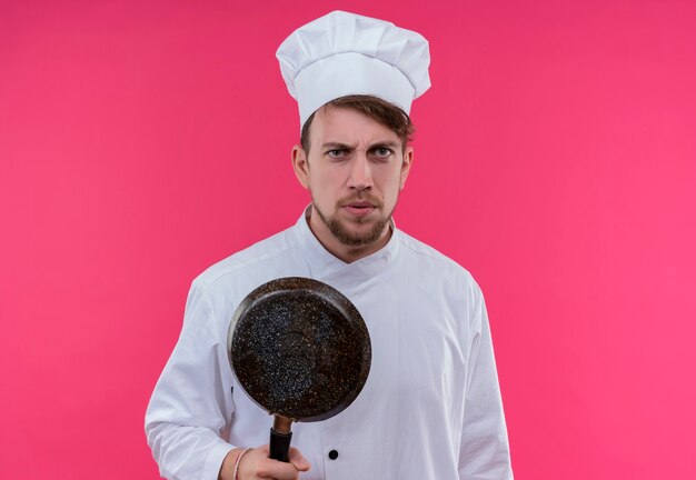 Un jeune chef barbu stressant homme en uniforme blanc tenant une poêle tout en regardant sur un mur rose