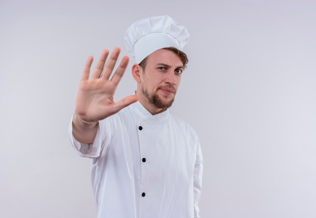 Un jeune chef barbu stressant homme vêtu d'un uniforme de cuisinière blanc et chapeau montrant le geste d'arrêt avec la main tout en regardant sur un mur blanc