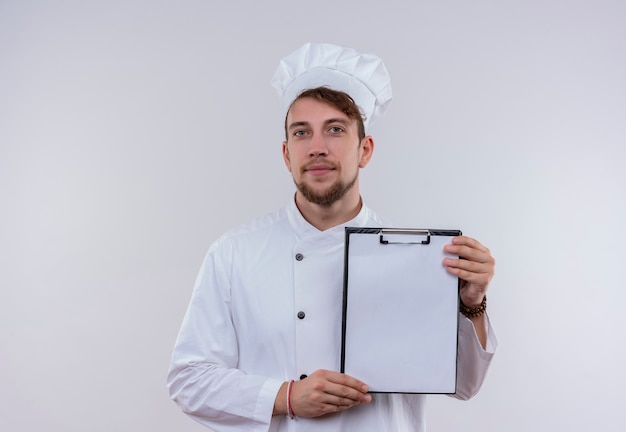 Un jeune chef barbu en uniforme blanc montrant un dossier vierge tout en regardant sur un mur blanc