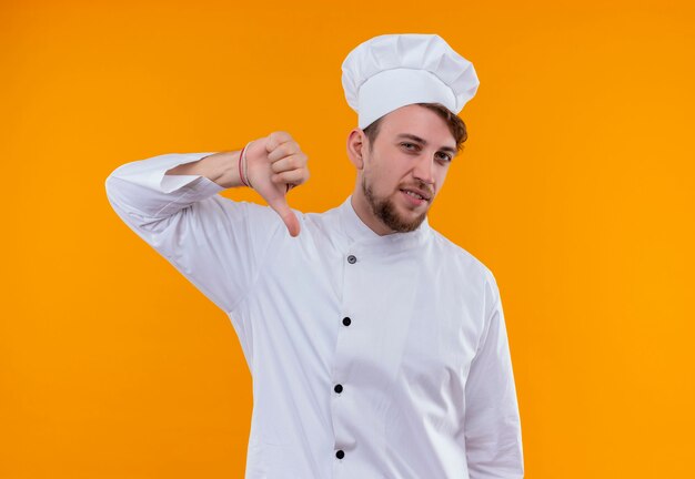 Un jeune chef barbu en uniforme blanc montrant les pouces vers le bas sur un mur orange