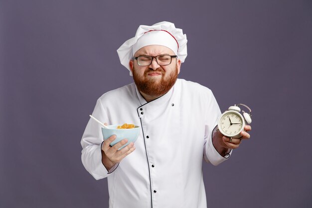 Jeune chef irrité portant des lunettes uniformes et une casquette tenant un réveil et un bol de pâtes macaronis avec une cuillère dedans avec les yeux fermés isolés sur fond violet