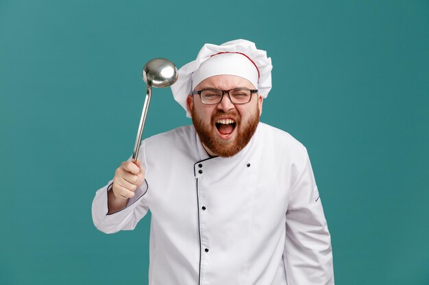 Jeune chef masculin furieux portant des lunettes uniformes et une casquette tenant une louche regardant la caméra criant fort isolé sur fond bleu