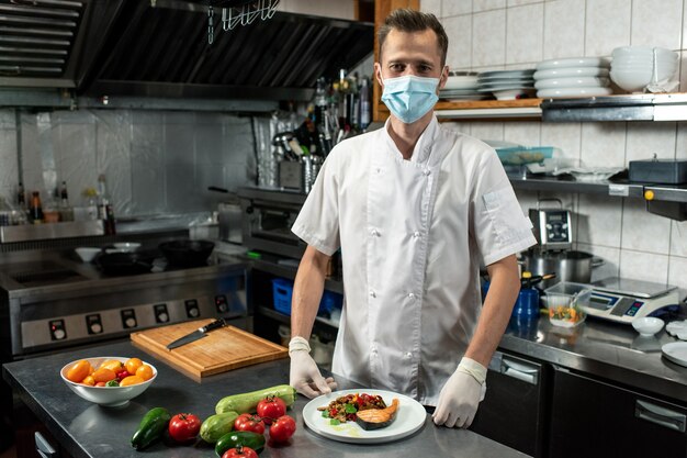 Jeune chef professionnel en uniforme blanc et masque de protection tenant du saumon frit avec des légumes hachés cuits à la vapeur sur une assiette dans la cuisine