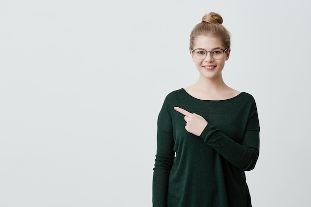 Jeune cliente ou étudiante gaie avec des cheveux blonds dans un chandail vert décontracté et des lunettes souriant largement, pointant l'index sur le mur de fond gris pour du texte ou des informations publicitaires