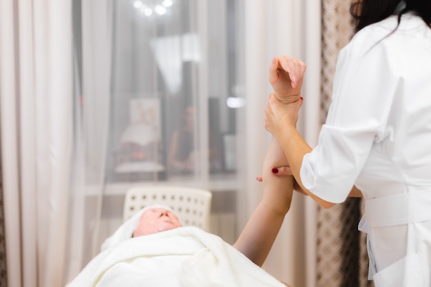 Une jeune cliente avec un masque d'alginate sur son visage se trouve sur la table de cosmétologie, l'esthéticienne fait un massage manuel des mains