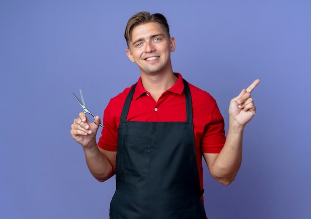 Jeune coiffeur blond souriant en uniforme tient des ciseaux et des points sur le côté