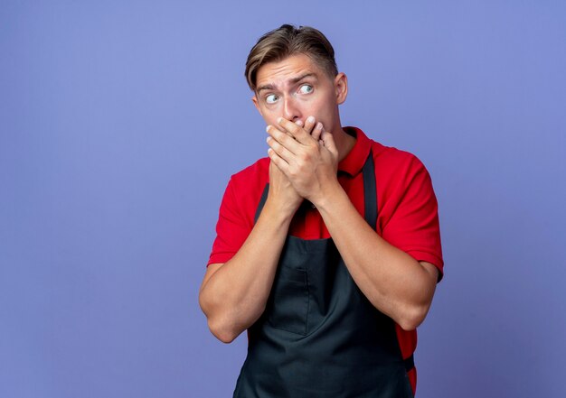 Jeune coiffeur homme blond choqué en uniforme met les mains sur la bouche en regardant côté isolé sur l'espace violet avec copie espace