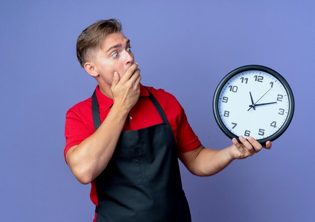 Jeune coiffeur homme blond choqué en uniforme tient et regarde l'horloge isolée sur l'espace violet avec copie espace