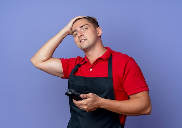 Jeune coiffeur homme blond confiant en uniforme met la main sur la tête tenant le peigne isolé sur l'espace violet avec copie espace