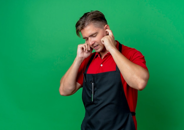 Jeune coiffeur homme blond ennuyé en uniforme bloque les oreilles avec les doigts isolés sur l'espace vert avec copie espace