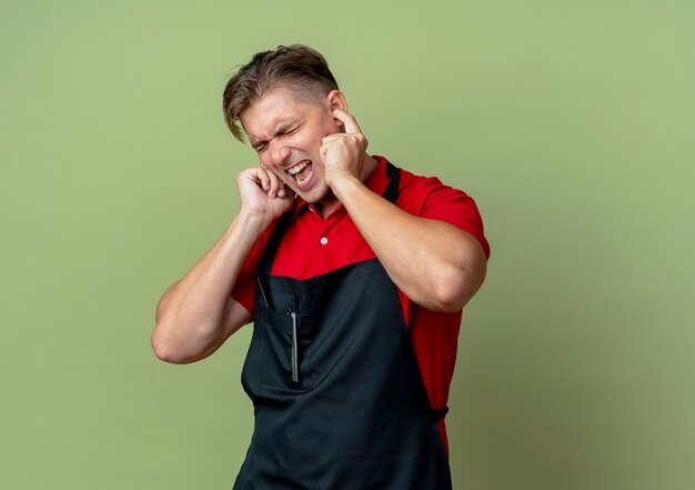 Jeune coiffeur homme blond ennuyé en uniforme bloque les oreilles avec les doigts isolés sur l'espace vert olive avec copie espace