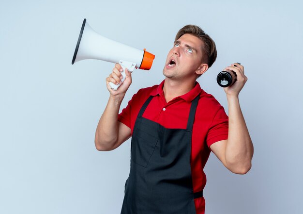 Jeune coiffeur homme blond ennuyé en uniforme détient haut-parleur et vaporisateur à la recherche d'isolement sur l'espace blanc avec copie espace