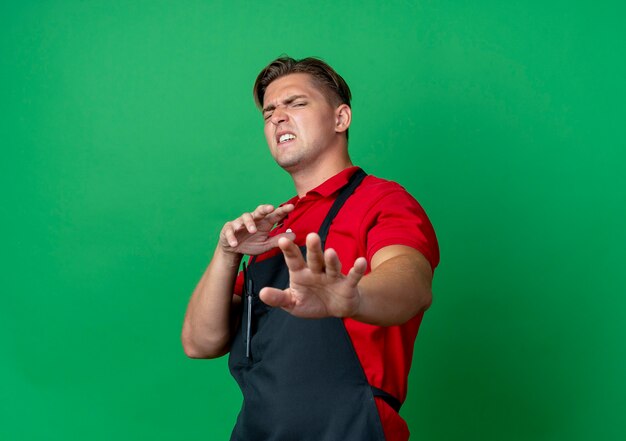 Jeune coiffeur homme blond ennuyé en uniforme fait semblant de pousser les mains étirées isolées sur l'espace vert avec copie espace