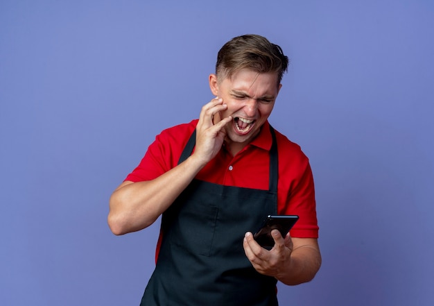 Jeune coiffeur homme blond ennuyé en uniforme met la main sur le visage en regardant téléphone isolé sur espace violet avec copie espace