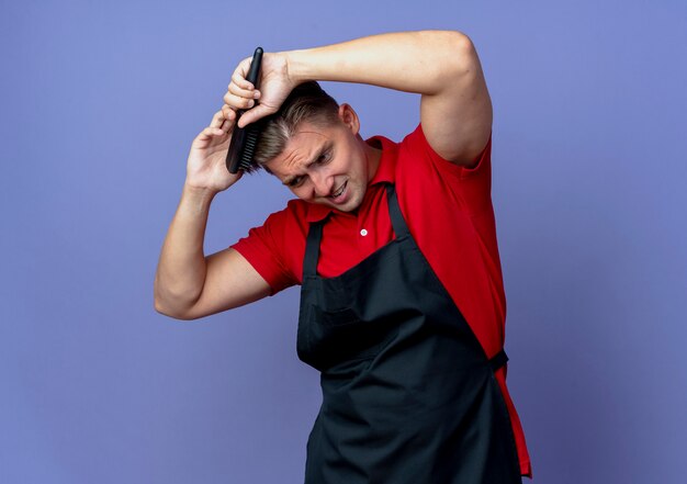 Jeune coiffeur homme blond ennuyé en uniforme peignant les cheveux regardant vers le bas isolé sur l'espace violet avec copie espace