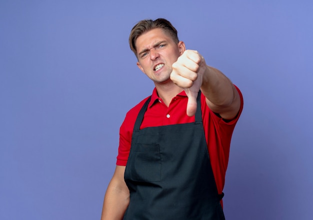 Jeune coiffeur homme blond ennuyé en uniforme pouces vers le bas isolé sur l'espace violet avec copie espace