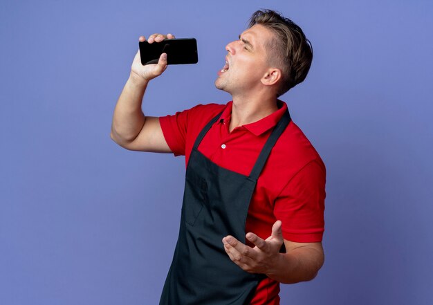 Jeune coiffeur homme blond ennuyé en uniforme se tient sur le côté tenant le téléphone et faisant semblant de chanter isolé sur l'espace violet avec copie espace