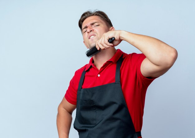 Jeune coiffeur homme blond ennuyé en uniforme tient le peigne sur le menton isolé sur un espace blanc avec copie espace