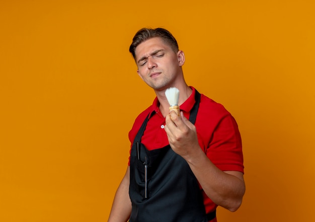 Jeune coiffeur homme blond ennuyé en uniforme tient et regarde le blaireau isolé sur l'espace orange avec copie espace