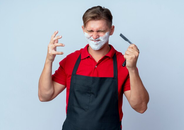 Jeune coiffeur homme blond ennuyé en uniforme visage enduit de mousse à raser tient un rasoir droit avec les yeux fermés isolé sur un espace blanc avec copie espace