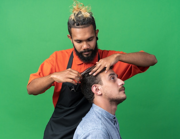 Jeune coiffeur masculin afro-américain concentré en uniforme faisant une coupe de cheveux pour son jeune client