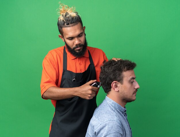 Jeune coiffeur masculin concentré en uniforme faisant une coupe de cheveux pour son jeune client isolé sur un mur vert