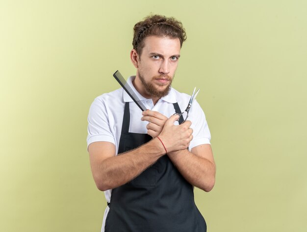 Jeune coiffeur masculin confiant portant l'uniforme tenant et traversant des sciccors avec un peigne isolé sur un mur vert olive