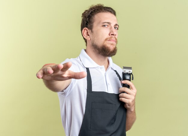 Jeune coiffeur masculin confiant en uniforme tenant une tondeuse à cheveux et tenant la main isolée sur un mur vert olive