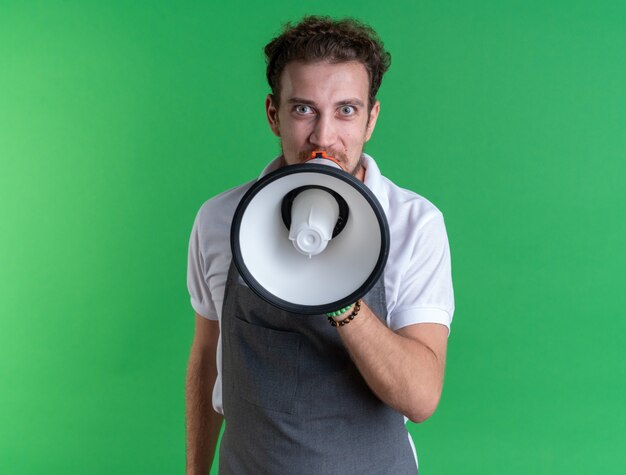 Un jeune coiffeur masculin excité en uniforme parle sur un haut-parleur isolé sur un mur vert